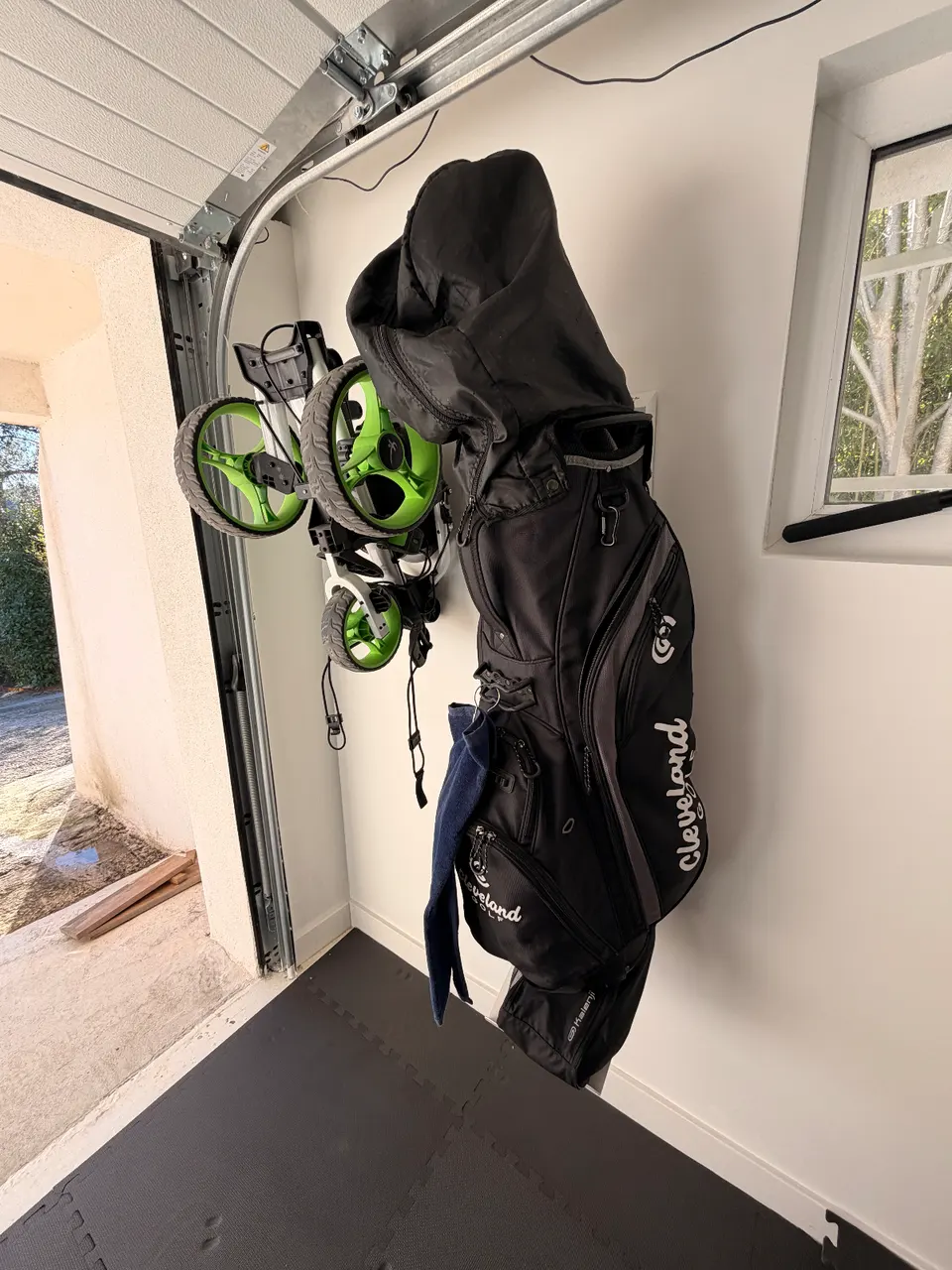 Sac de golf noir suspendu à un mur, avec un support de caddy et un coin de garage visible.